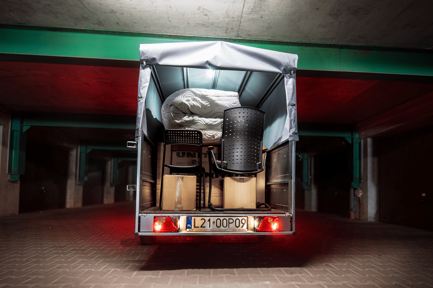 Vue arrière de l'intérieur d'une remorque Unitrailer avec bâche haute ouverte, chargée d'objets de déménagement comprenant deux chaises noires, des cartons et un matelas enroulé, stationnée dans un parking souterrain.