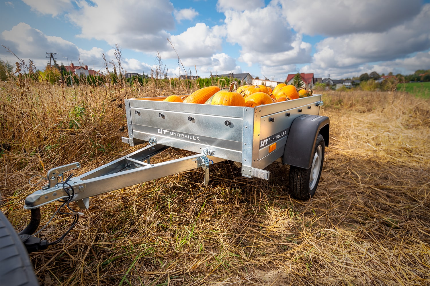 Remorque mono-axe Unitrailer remplie de citrouilles oranges, stationnée dans un champ d'herbe séchée sous un ciel nuageux.