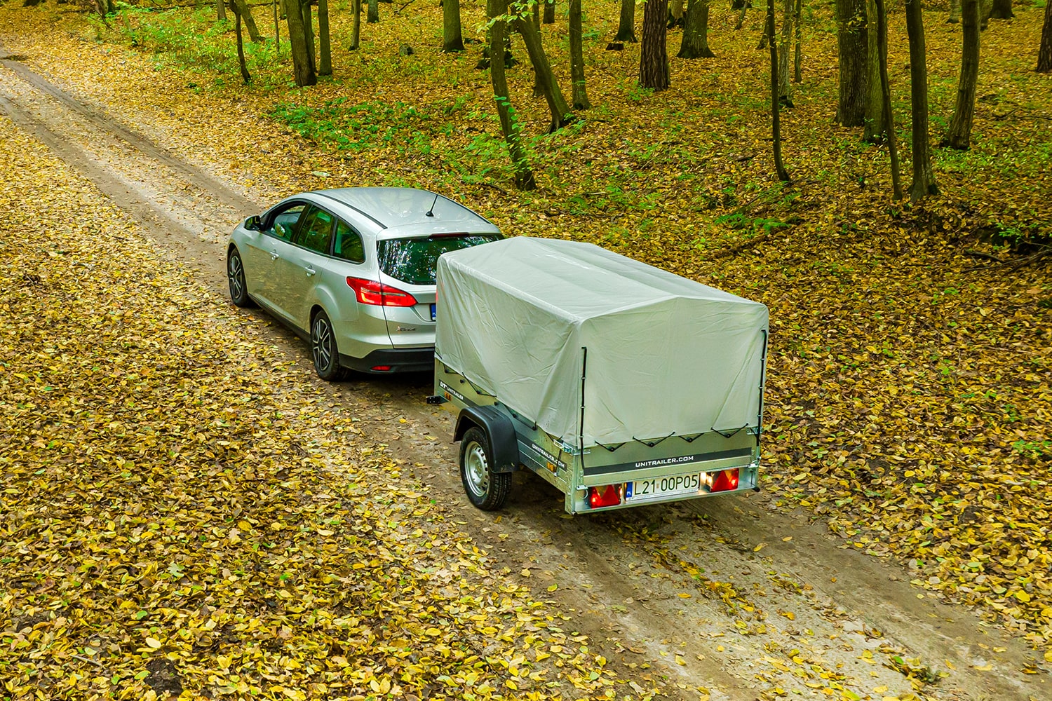 Vue aérienne d'une voiture break grise tractant une remorque Unitrailer équipée d'une bâche haute grise sur un chemin forestier recouvert de feuilles d'automne.